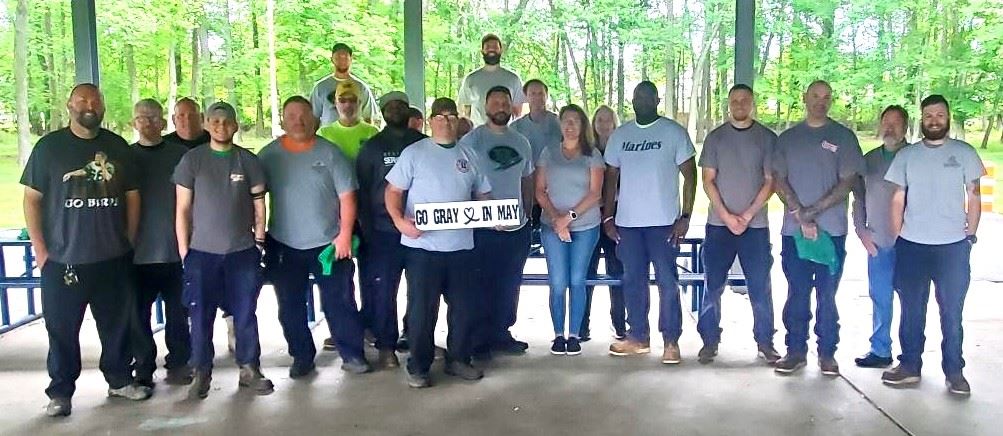 Group of about 16 adults standing outdoors in a wooded park; some wear matching shirts on a patio.